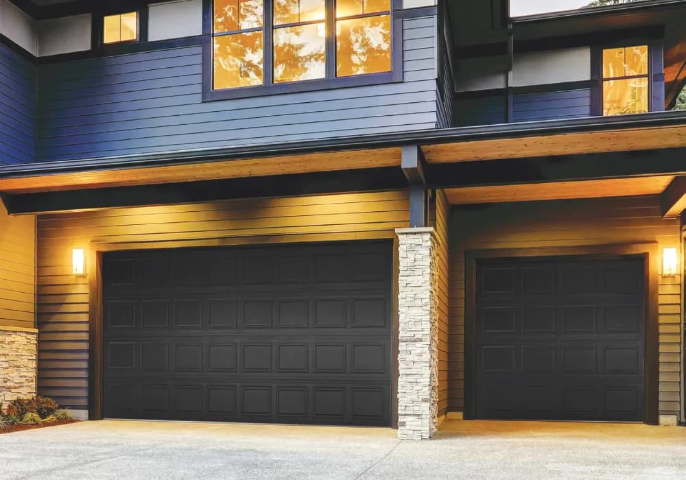 Black raised-panel garage doors under warm evening lighting on a modern home